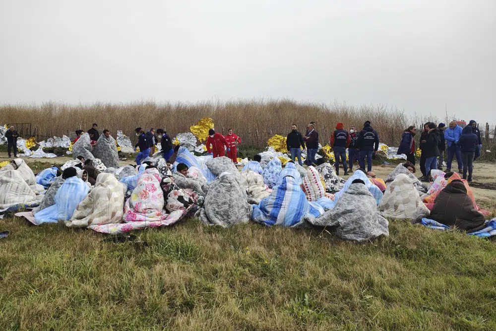 Migrantes rescatados se sientan cubiertos con mantas en una playa cerca de Cutro, en el sur de Italia, el domingo 26 de febrero de 2023. Los funcionarios de rescate dicen que un número indeterminado de migrantes ha muerto y que decenas han sido rescatados después de que su bote se rompiera en el sur de Italia.  (Antonino Durso/LaPresse vía AP)