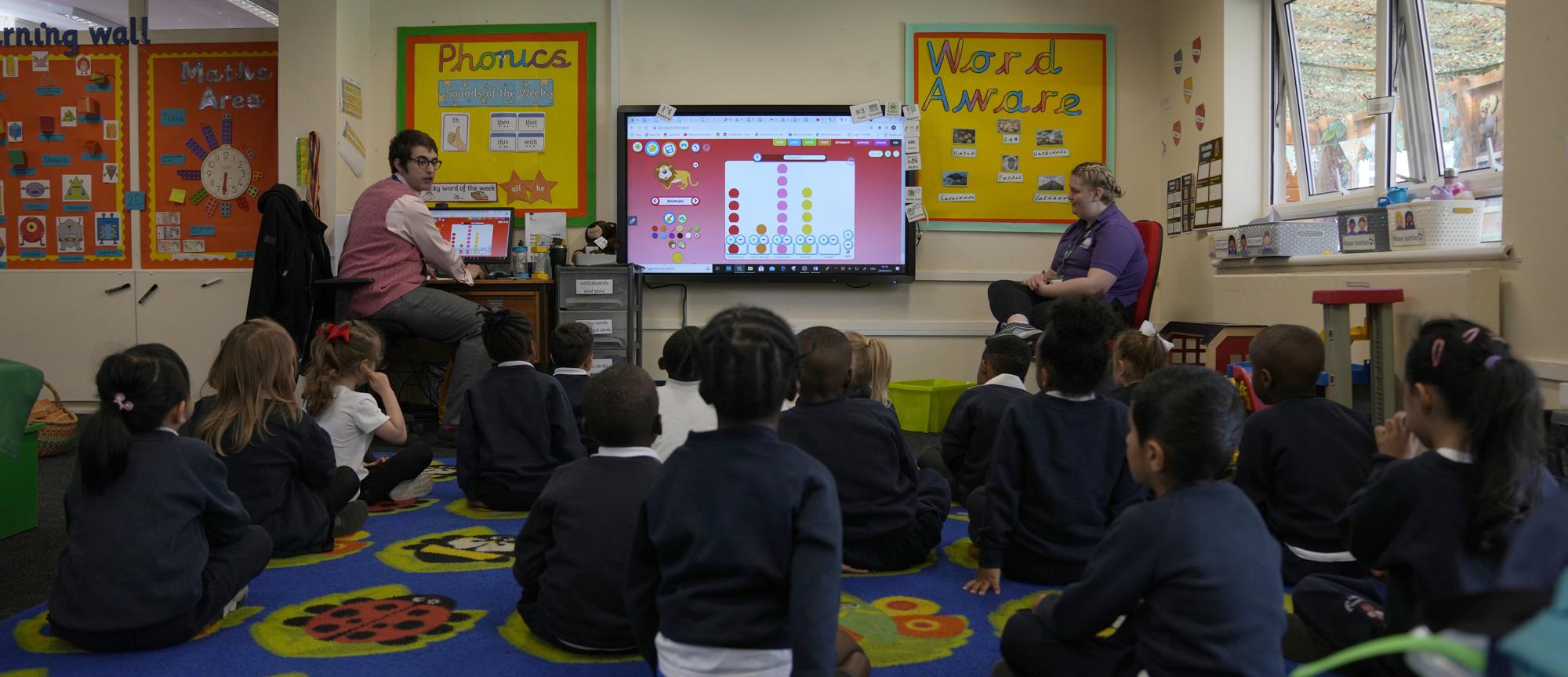 Alex Dickerson the reception class teacher, left leads the class at the Holy Family Catholic Primary School in Greenwich, London, Monday, May 24, 2021. Holy Family, like schools across Britain, is racing to offset the disruption caused by COVID-19, which has hit kids from low-income and ethnic minority families hardest. (AP Photo/Alastair Grant)