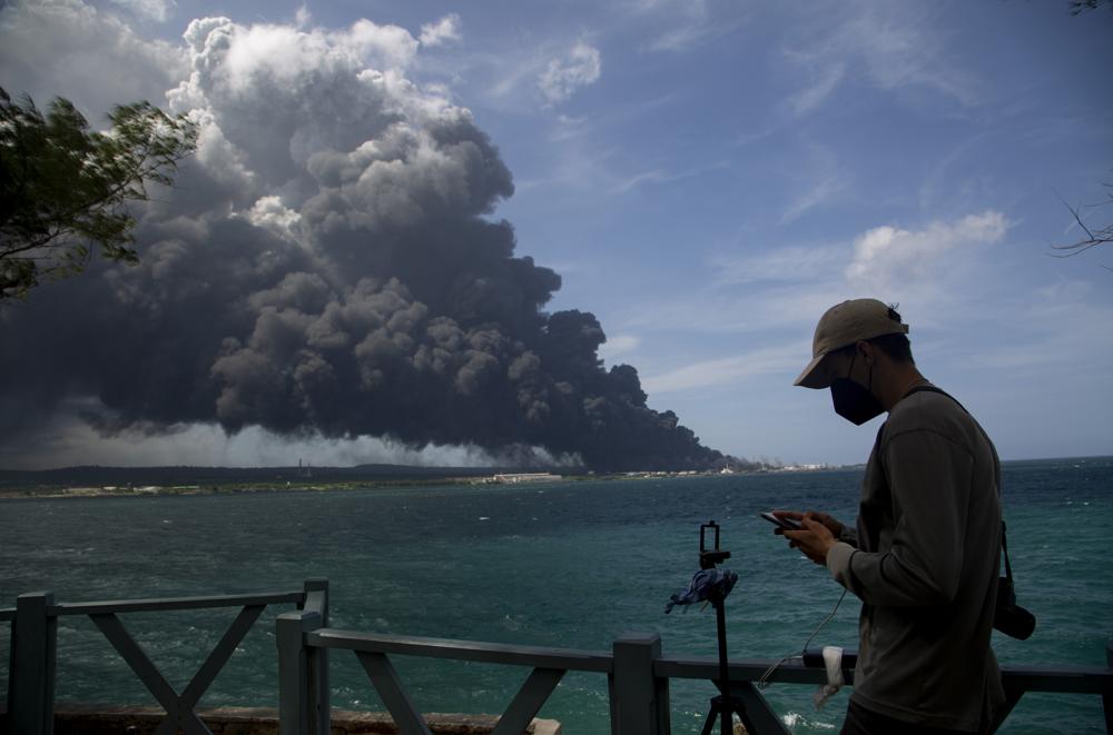 A man types on his cellphone near a huge plume of smoke rising from the Matanzas Supertanker Base as firefighters work to quell the blaze which began during a thunderstorm in Matanzas, Cuba, Monday, Aug. 8, 2022. Cuban authorities say lightning struck a crude oil storage tank at the base, sparking a fire that sparked four explosions. (AP Photo/Ismael Francisco)