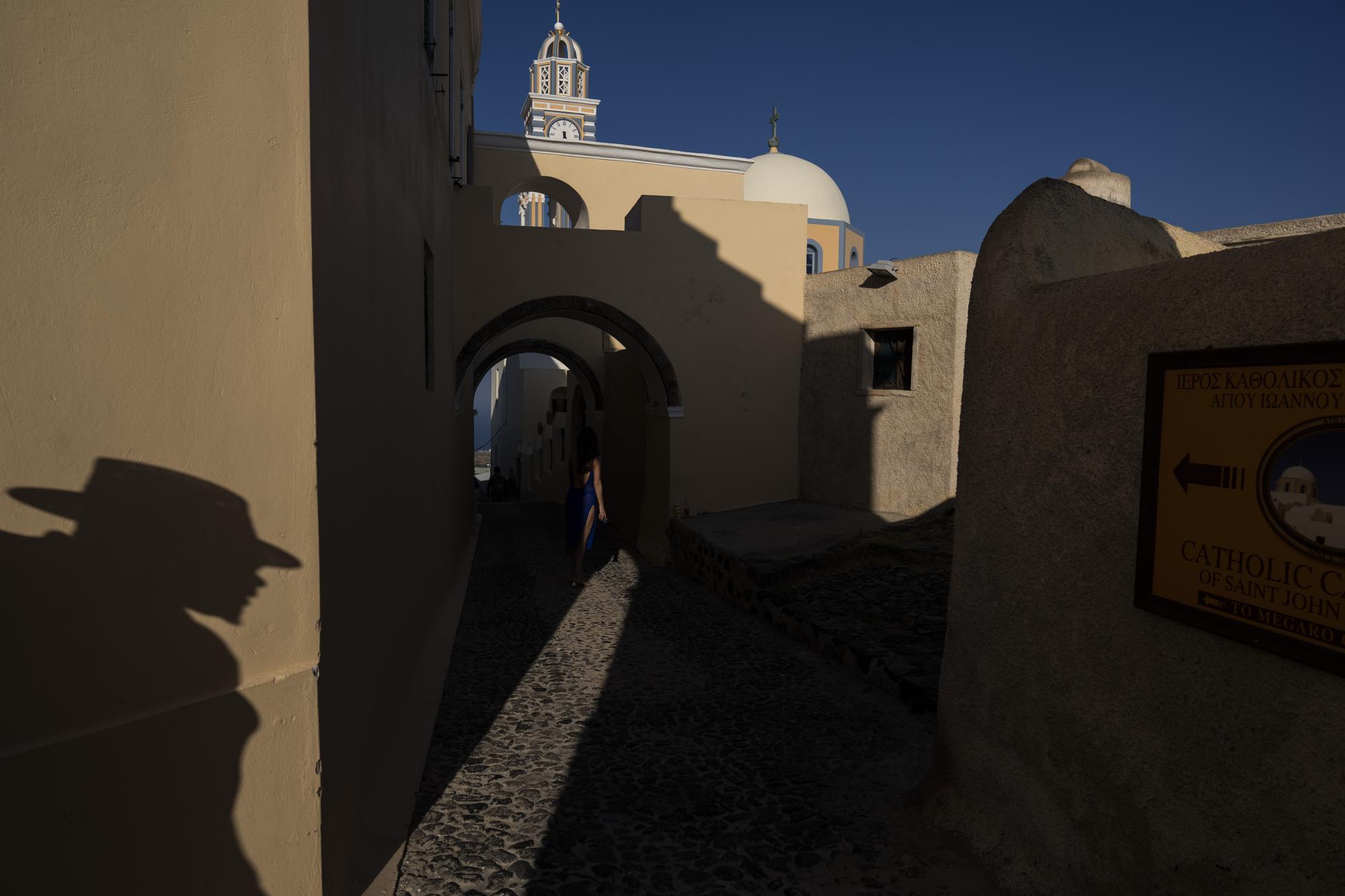 Una mujer posa en un callejón entre la Catedral de San Juan Bautista, a la derecha, y el Monasterio de Santa Catalina en la isla griega de Santorini el miércoles 15 de junio de 2022. Dentro del convento, 13 monjas de clausura dedican sus vidas a oración.  (Foto AP/Petros Giannakouris)