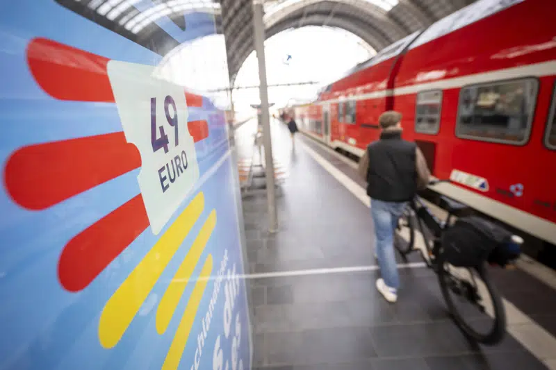 A man passes by at the main train station on his bicycle as an advertising for the Deutschlandticket (Germany Ticket) is seen at left, in Frankfurt, Germany, Monday, May 1, 2023. Public transit companies in Germany say more than three million people have already snapped up a new ticket being launched Monday that allows them to use all local and regional trains, buses and metros across the country for 49 euros ($53.90) a month. (Sebastian Gollnow/dpa via AP)