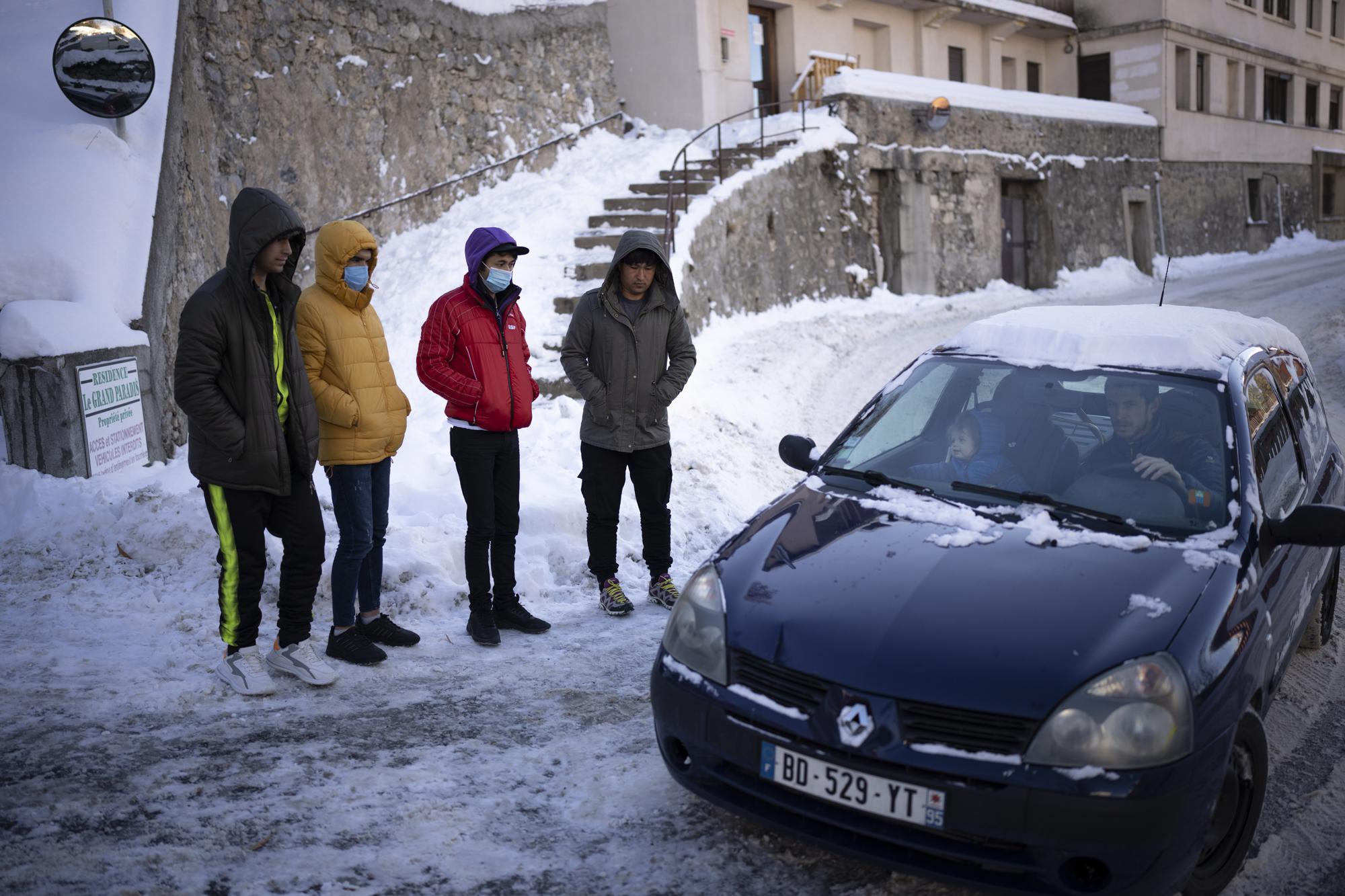 A family drives past Afghan migrants waiting outside a migrant refuge to be taken to have a PCR test in Briancon, France, Saturday, Dec. 11, 2021. (AP Photo/Daniel Cole)