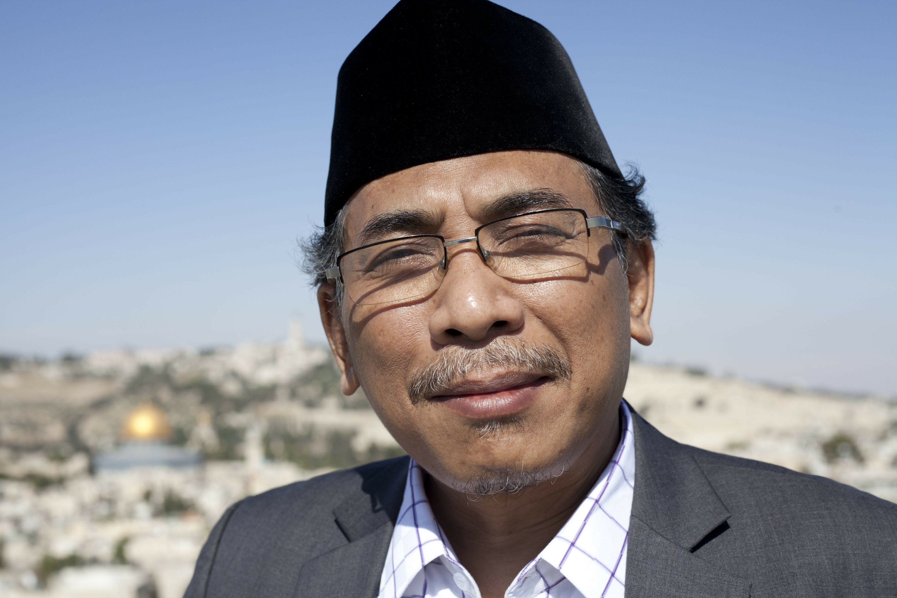  A Muslim spiritual advisor, wearing a black cap and glasses, smiles in front of a blurred background of a cityscape.