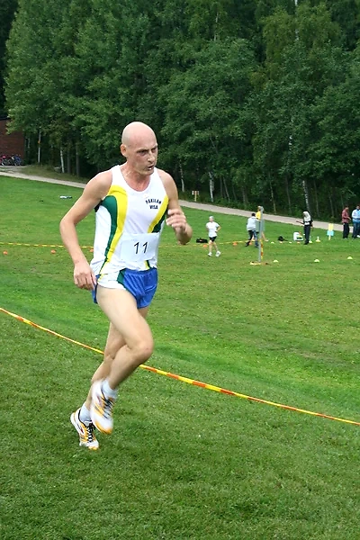 The cross-country runner with number 11 is running on the grassy field, with a forest in the background.