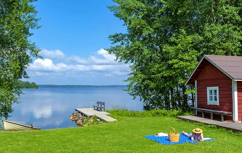 A beautiful lake landscape with a red cabin, a dock, and a person resting on the lawn.