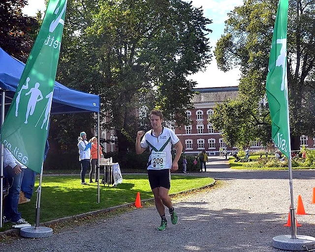 Runner in the Årungen Rundt race with bib number 209, cheering towards the finish line. Green flags and spectators in the background.