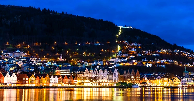 Night picture of the city of Bergen, glowing buildings along the water and up the mountainside.