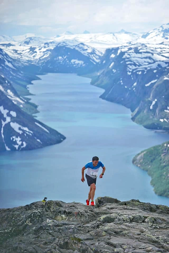 En løper klatrer en steinete sti med fjell og innsjø i bakgrunnen.