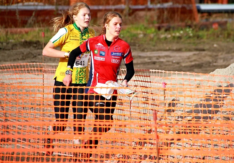 Two women running orienteering, with a map in hand, behind an orange fence.