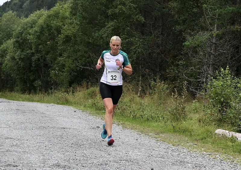 Løper i terrengløp. Kvinne i startnummer 32 løper på grusvei med skog i bakgrunnen.