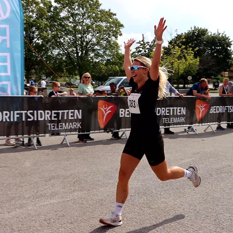 The runner cheers at the finish line with arms raised. The audience watches from the sidelines.