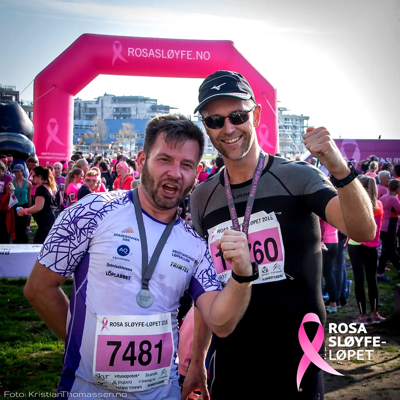 Men celebrate the Pink Ribbon race, with medals and bib numbers, in front of the Pink Ribbon arch.