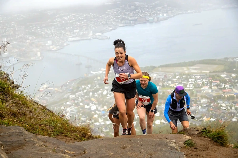 Runners climb a steep mountain trail in an endurance race with a city in the background.
