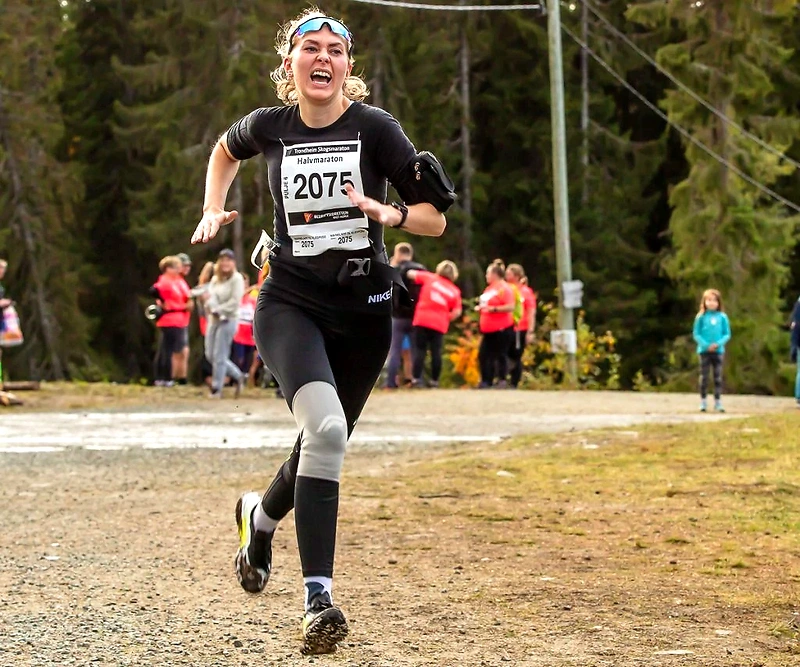 Female runner in Trondheim Forest Marathon, wearing a bib number, running in a forest environment.