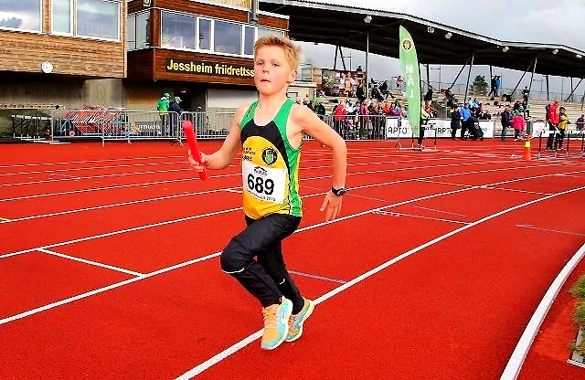 Young runner on the track, holding a relay baton. Audience in the background.