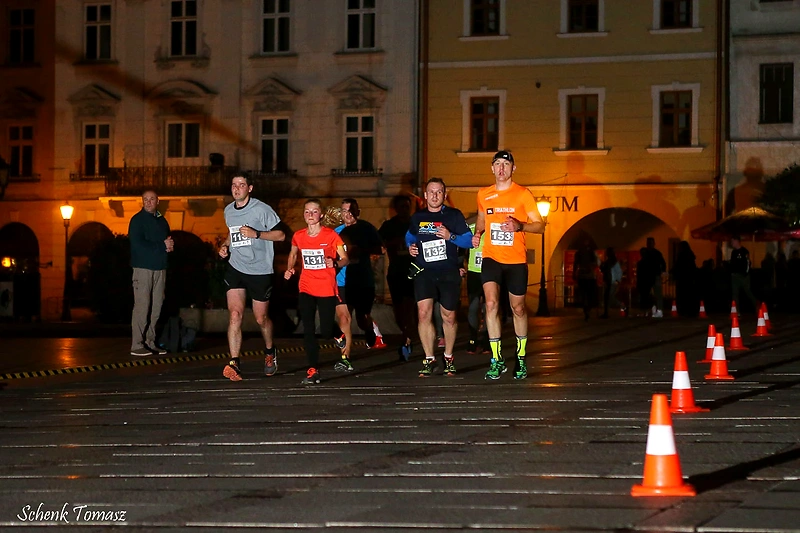 Biegacze podczas nocnego biegu w mieście, widoczne numery startowe, oświetlenie i pachołki.