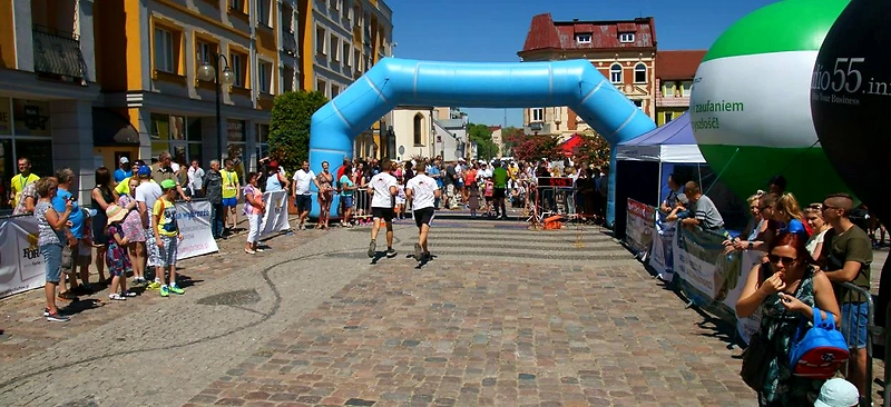 Biegacze na starcie biegu ulicznego, z widocznymi kibicami i bramą startową.
