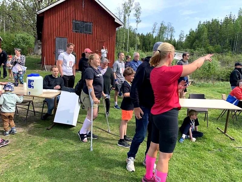 Löpare och åskådare vid ett terränglopp. Ett bord med vatten och en röd stuga i bakgrunden.