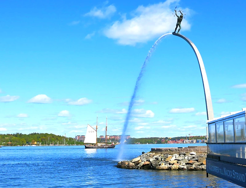 Utsikt över Nacka Strand med vattenspel och segelbåt. Välkommen till Nacka Strand.