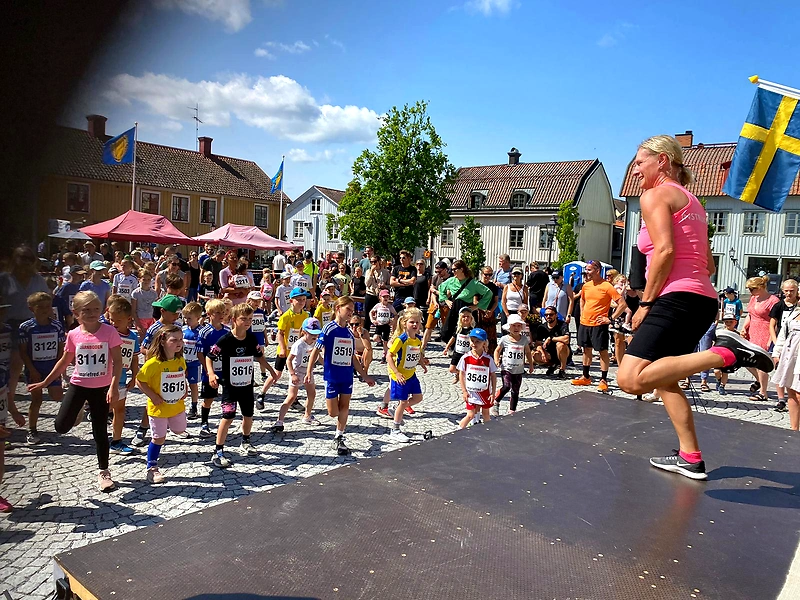 Children are running in a race with spectators and flags in the background.