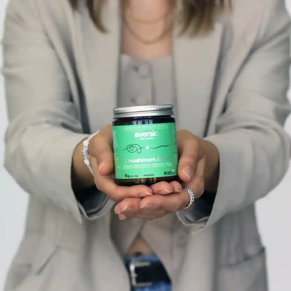 A person in a beige blazer holding the green jar of Eversio functional mushroom blend supplement. A person in a beige blazer holding the green jar of Eversio functional mushroom blend supplement.