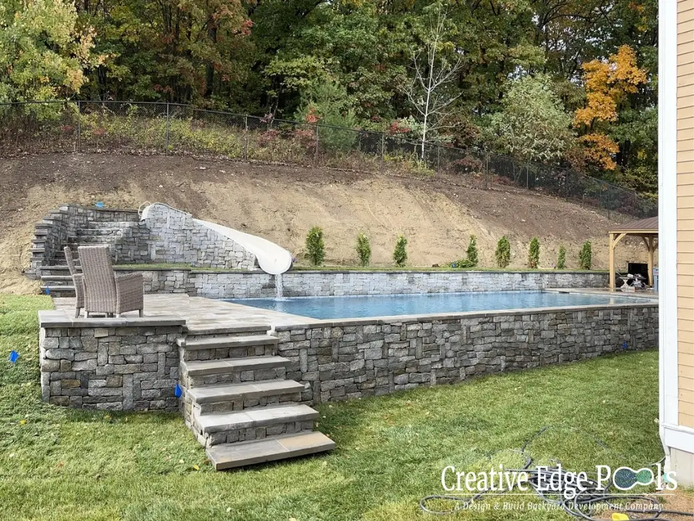 A luxury pool built on a sloping New England backyard lot with stone retaining walls - North Shore pool builders
