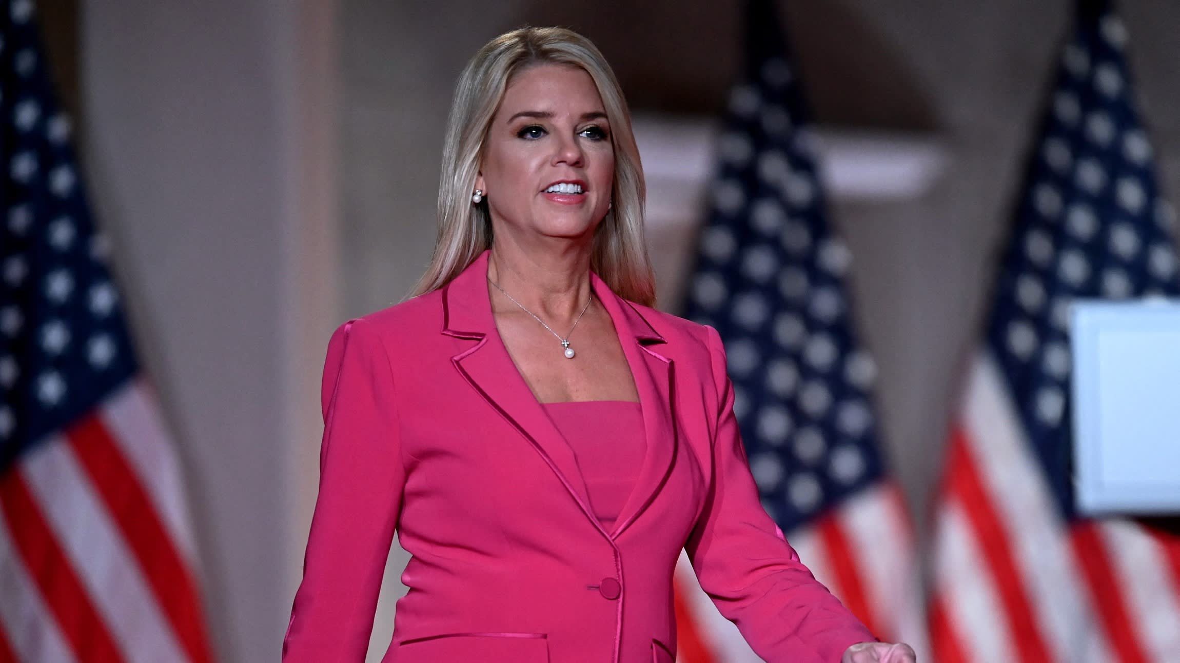 Pam Bondi caminando con traje rosa ante banderas estadounidenses