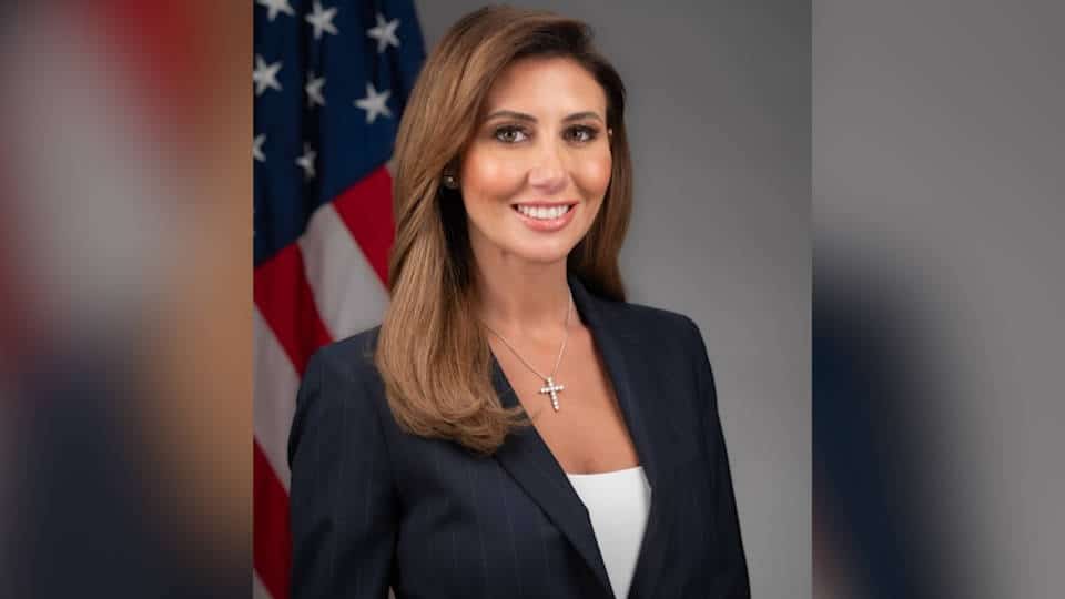 Alina Habba sonriente con traje oscuro y bandera estadounidense de fondo