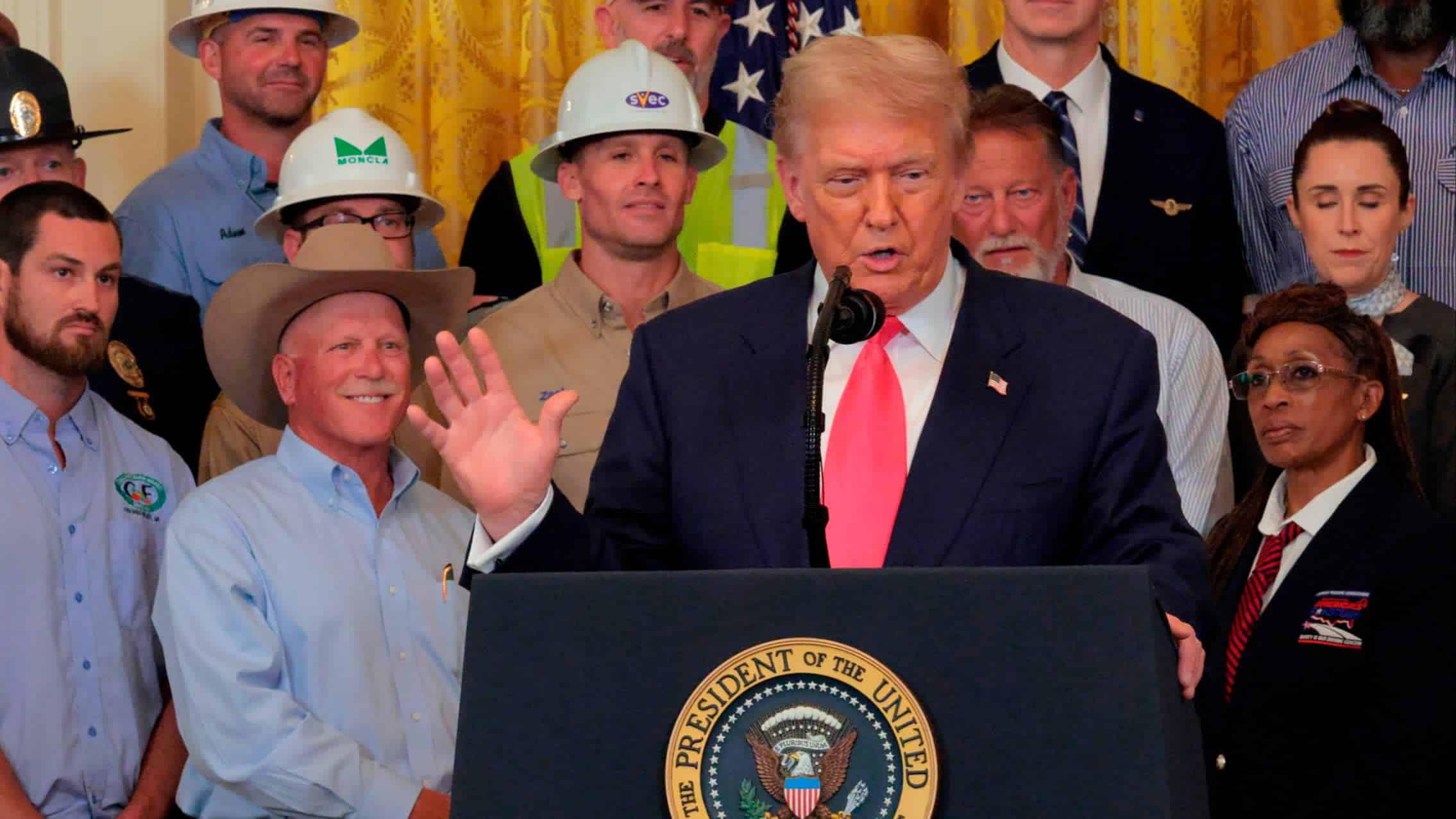 Donald Trump hablando en un podio con el sello presidencial, rodeado de asistentes con cascos de construcci&oacute;n