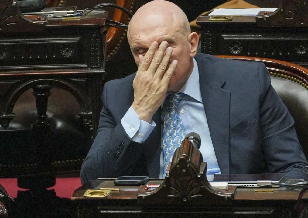 Hombre con cabeza rapada en entorno legislativo mostrando frustración durante reunión de comisión