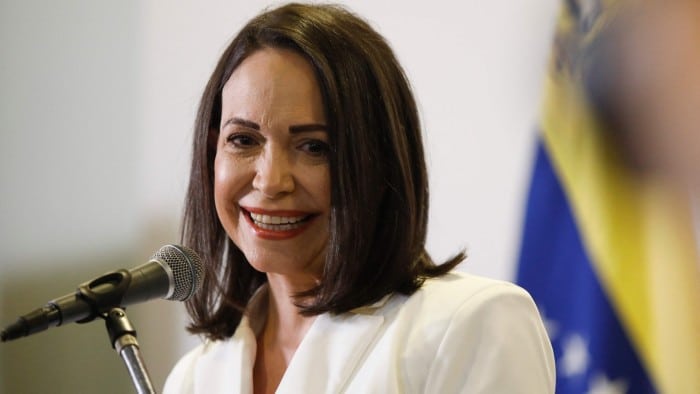 Mar&iacute;a Corina Machado hablando por un micr&oacute;fono con la bandera de Venezuela al fondo