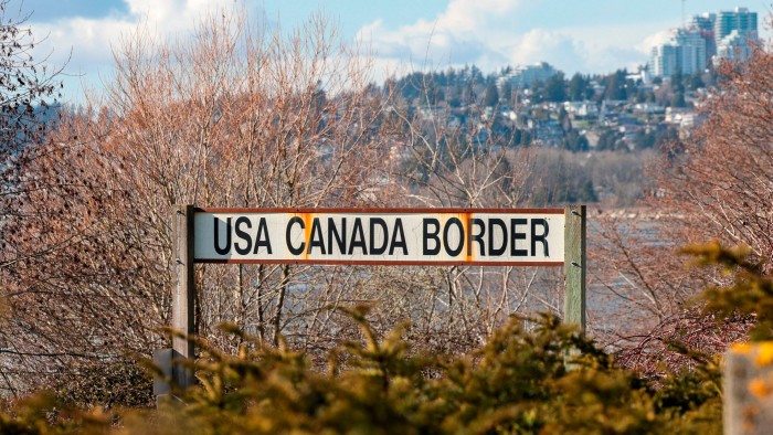 Letrero de la frontera entre Estados Unidos y Canad&aacute; con &aacute;rboles y edificios lejanos