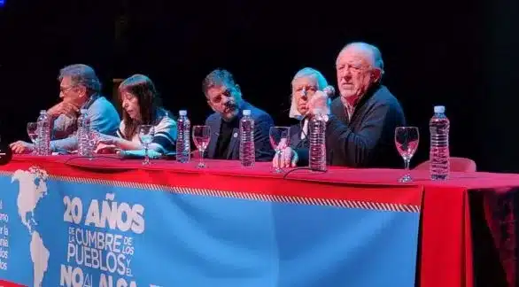 Mesa con banner que dice '20 A&Ntilde;OS DE CUMBRE DE LOS PUEBLOS Y EL NO AL ALCA'