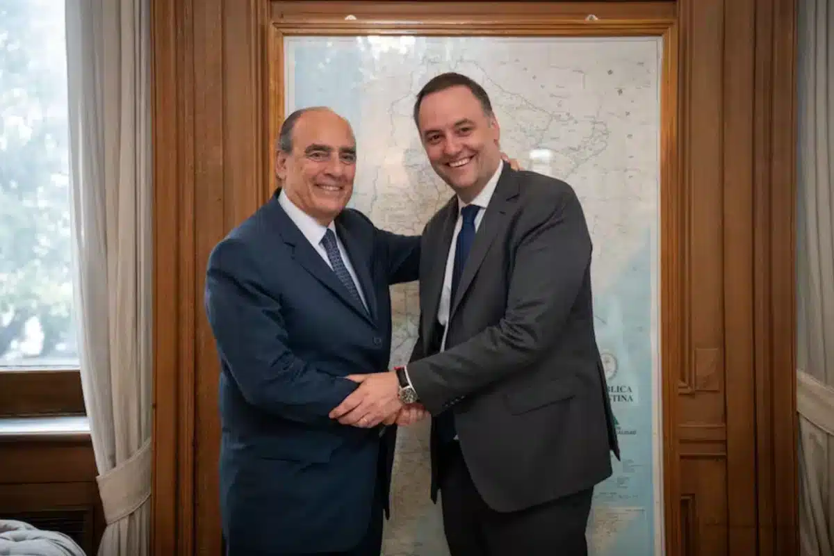 Manuel Adorni y Guillermo Francos sonriendo y d&aacute;ndose la mano en una oficina con un mapa de Argentina de fondo