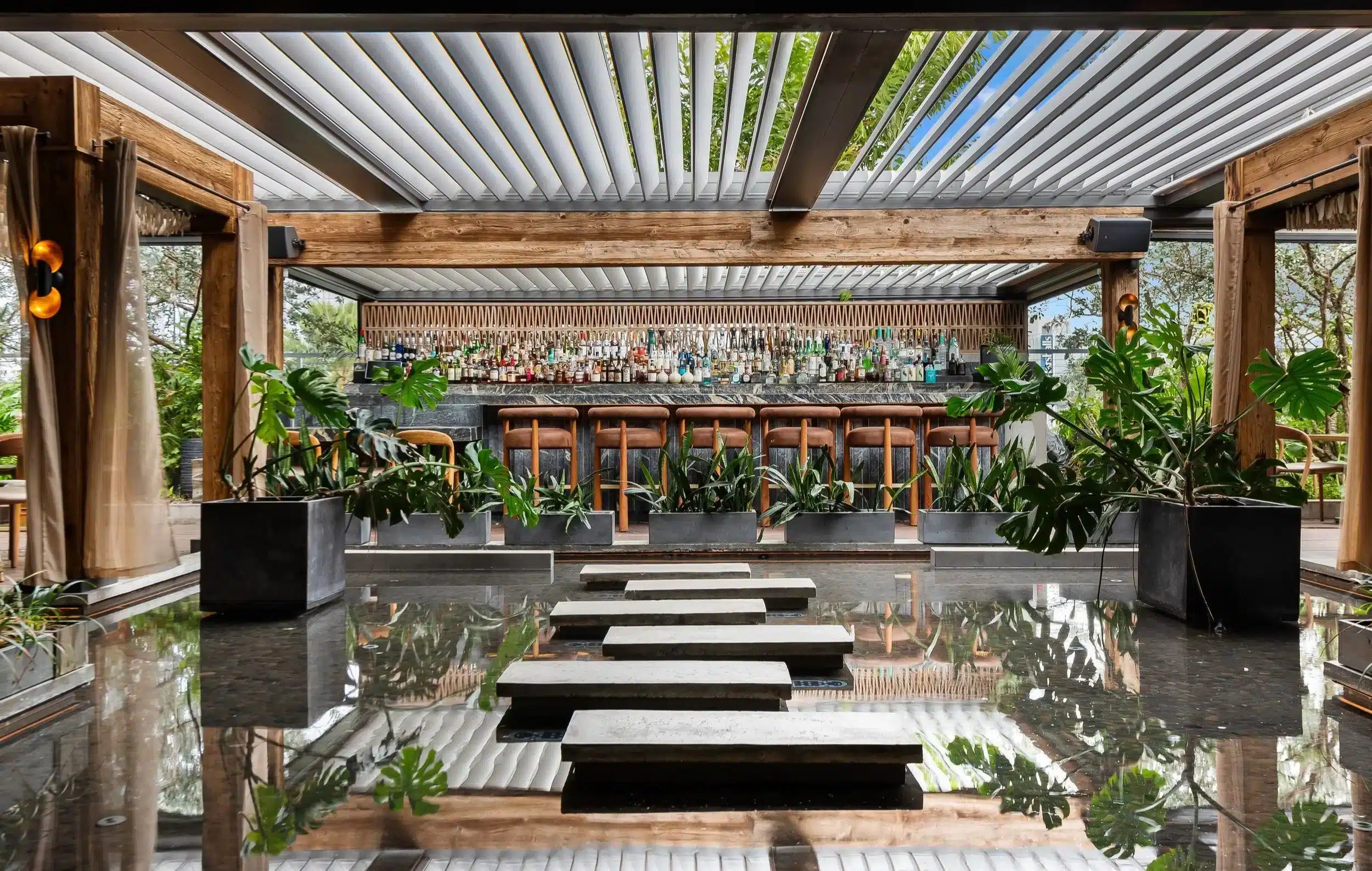 Interior luminoso del restaurante Mila con bar de madera oscura, estantes de botellas y plantas decorativas