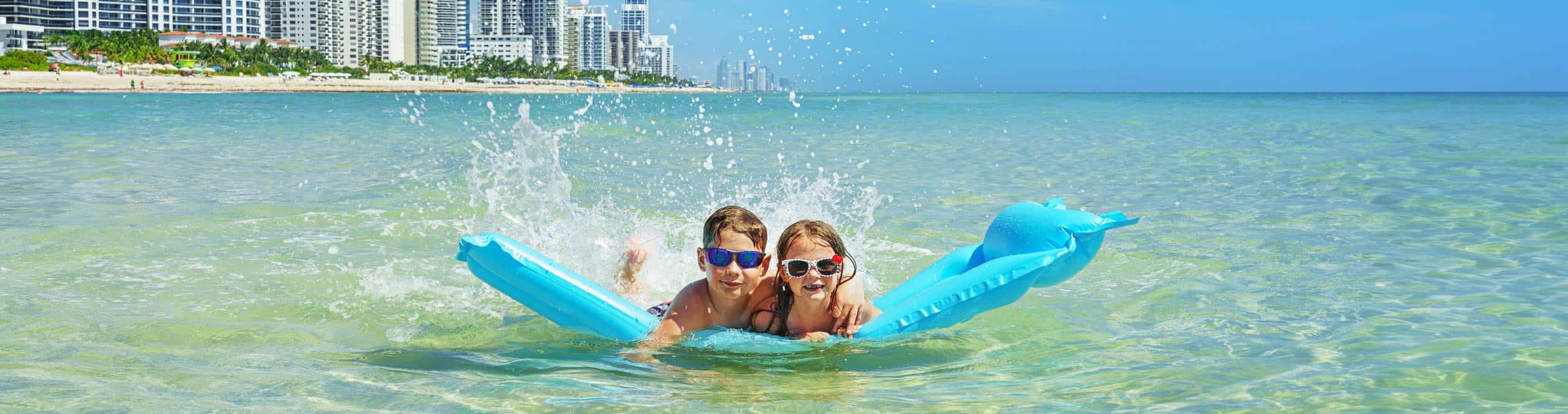 Escena genérica de un día soleado en el agua, con niños jugando con flotadores cerca de edificios altos, representativa de las actividades acuáticas en Miami