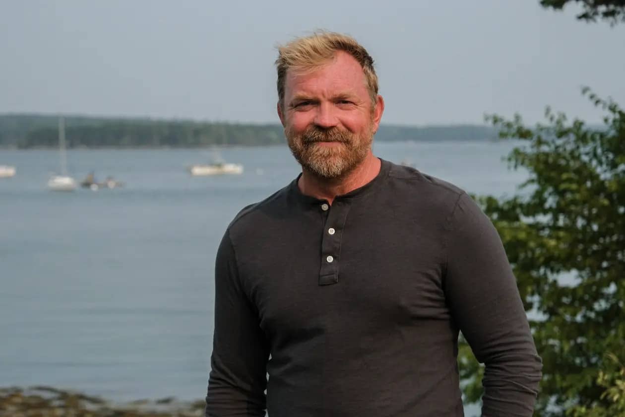 Graham Platner, ostricultor de Maine, de pie frente a un paisaje costero con barcos al fondo