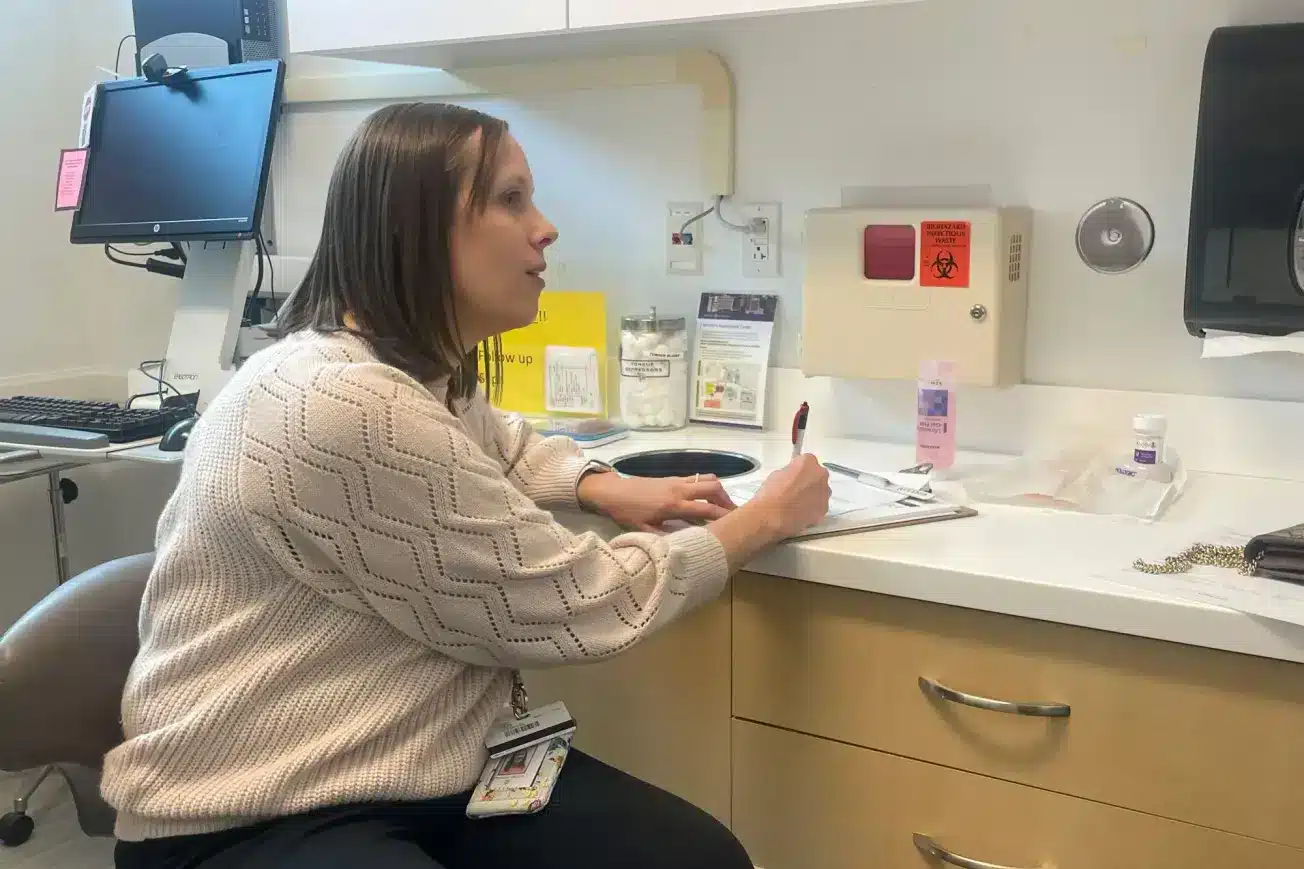 Una mujer sentada en un consultorio médico, tomando notas, mientras una computadora y suministros médicos se ven al fondo