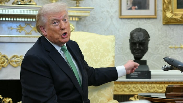 Individuo gesticulando en un entorno formal, posiblemente la Oficina Oval, con un busto detrás