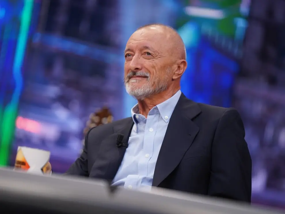 Arturo Pérez-Reverte sonriente en un set de televisión sosteniendo una taza