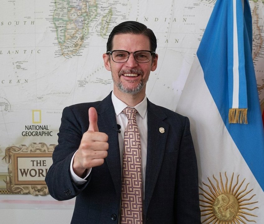 Sabino Vaca Narvaja sonriente con pulgar arriba frente a un mapa del mundo y la bandera argentina