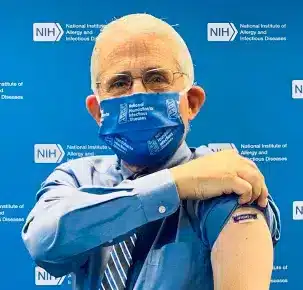 Persona con gafas y mascarilla azul mostrando una vacuna en el brazo con el logo del NIH de fondo