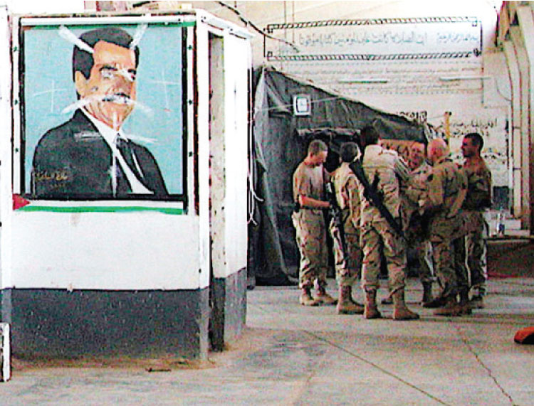 جنود أميركيون عند جدارية مشوهة لصدام حسين في معتقل بغداد المركزي، سجن أبو غريب سابقًا – (المصدر)