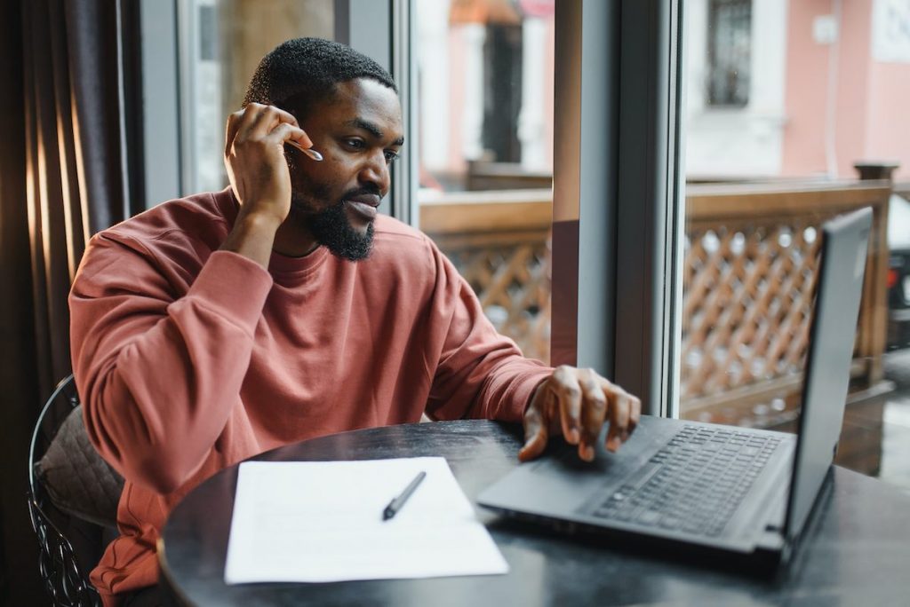 Afro american talks video chat on computer sits in cafe with cup of coffee. Black man calls on laptop on video link and speaks. He wears in shirt and suit jacket. Video link online connection.