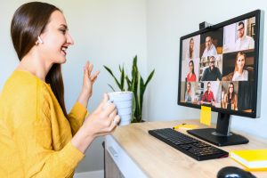 An employee smiles and waves to their remote team via a video call on their computer while holding a coffee cup