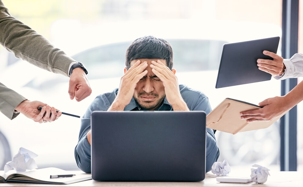 Man looking stressed holding his head with hands holding devices surrounding  him

