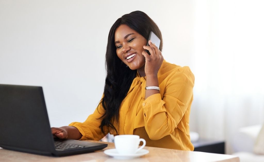 Smiling woman answering a call on a mobile phone, sitting at a desk with a laptop.