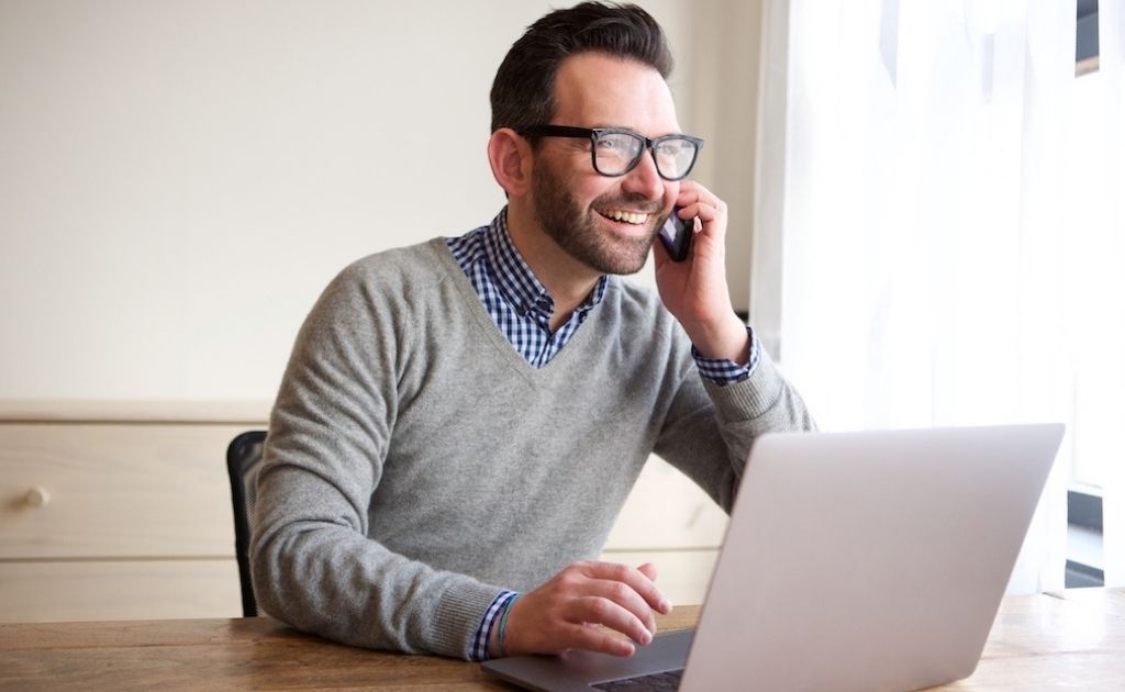 Smiling man on a mobile phone, sitting at a desk with a laptop.