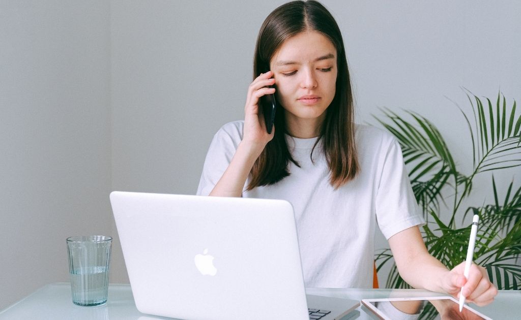 Woman on a mobile phone dealing with an angry caller, takes notes on a notepad in front on a laptop.