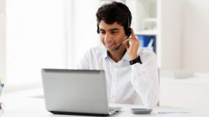 Smiling man answering business calls using a headset, sitting in front of a laptop